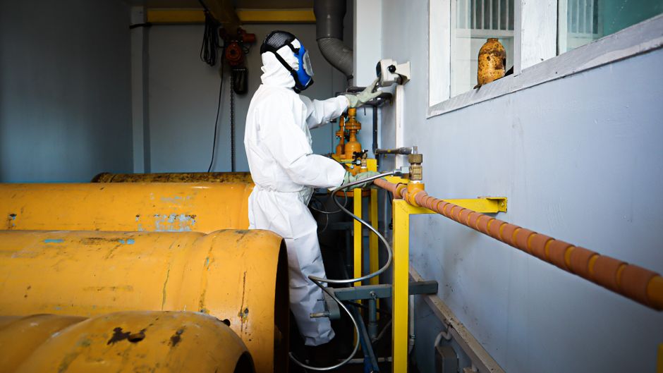 A worker in a white protective suit, gloves, and a face shield is operating machinery in an industrial setting. The worker is standing on a concrete floor beside large, yellow metal rollers with a tex