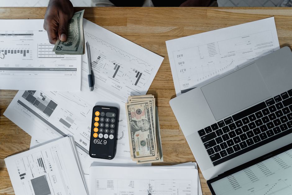 A wooden desk workspace featuring various documents, financial charts, and spreadsheets spread out across the surface. A person's dark-skinned hand is visible holding a twenty-dollar bill, with additi
