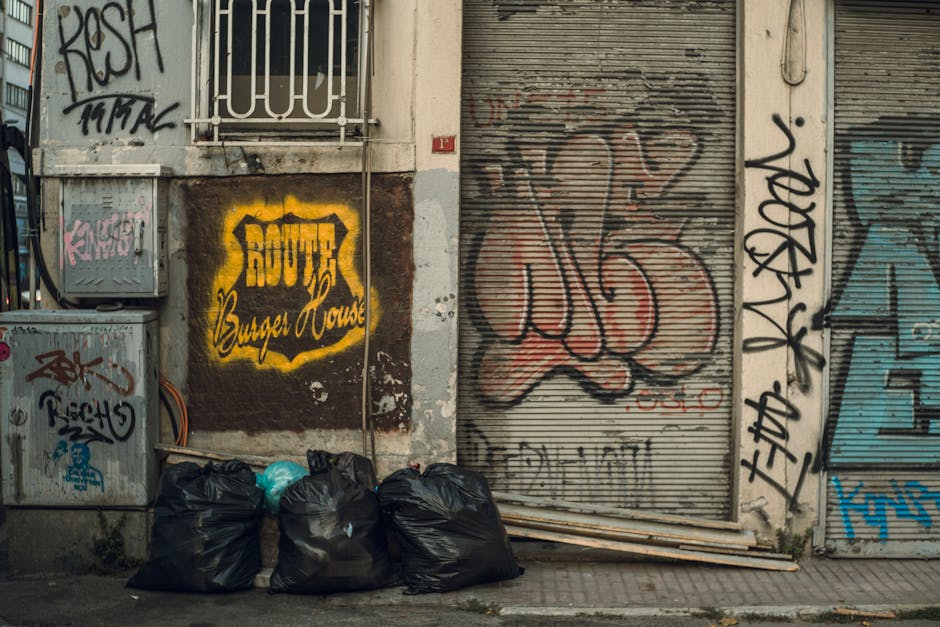 A waste collection operative, dressed in a green uniform with reflective orange strips on the trousers, stands beside a white and red rubbish truck on an urban street lined with green trees. The truck
